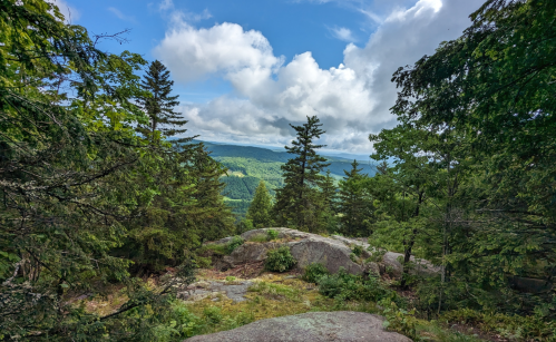 A scenic view from a rocky outcrop, surrounded by trees, overlooking a lush green valley under a partly cloudy sky.