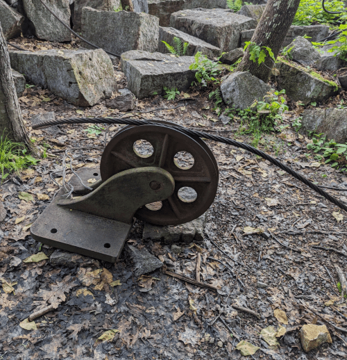 A rusty pulley on a rocky forest floor, surrounded by large stones and greenery.