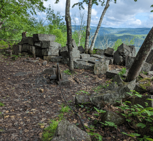 A rocky clearing in a forest with stacked stones, trees, and a distant view of hills under a cloudy sky.