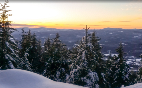 Snow-covered trees in the foreground with a sunset over distant mountains and valleys in a winter landscape.