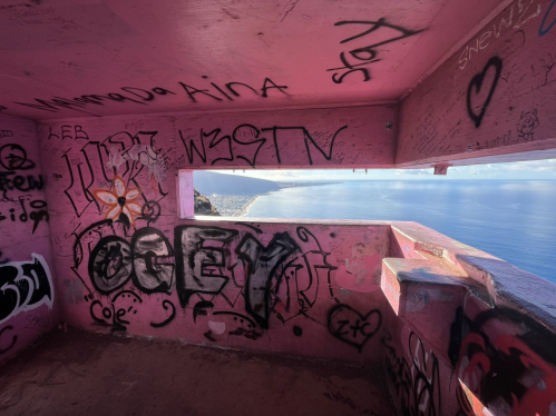 Interior of a pink-walled lookout tower covered in graffiti, with a view of the ocean through a window.