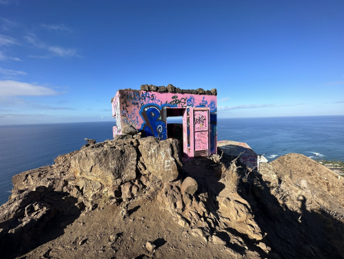 A colorful, graffiti-covered structure on a rocky hilltop overlooking the ocean under a clear blue sky.