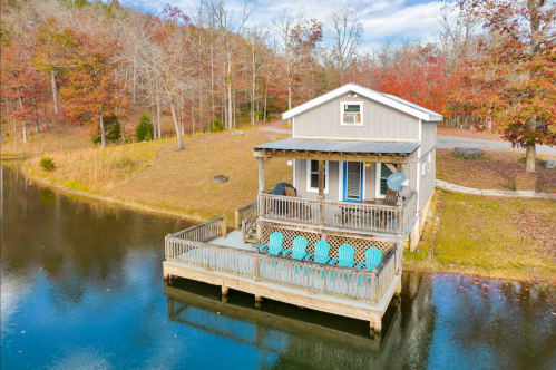 A cozy lakeside cabin with a deck and chairs, surrounded by autumn trees and calm water.