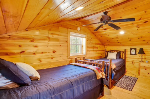 Cozy wooden bedroom with two beds, a ceiling fan, and a window, featuring warm wood paneling and soft lighting.