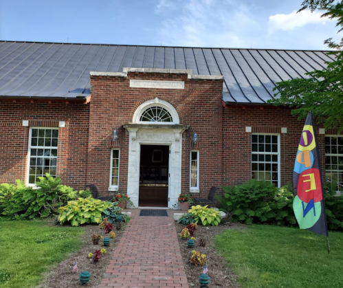 A brick building with a large entrance, surrounded by greenery and a colorful "OPEN" flag in the foreground.