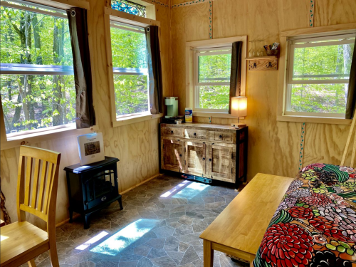 Cozy interior of a cabin with wooden walls, a table, chair, and windows overlooking a green forest.