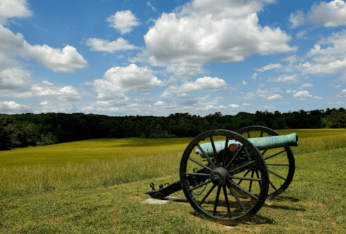 A historic cannon on a grassy field under a blue sky with fluffy clouds and trees in the background.