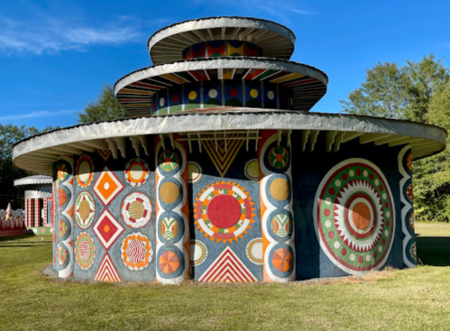 Colorful, patterned circular building with multiple tiers, surrounded by green grass and trees under a clear blue sky.