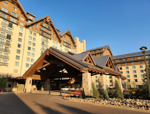 A large hotel entrance with a wooden canopy, stone pillars, and a backdrop of a multi-story building under a clear blue sky.