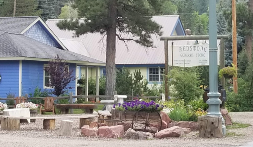 A quaint general store surrounded by greenery, flowers, and rustic seating in a peaceful rural setting.