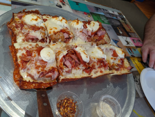 A large, cheesy pizza topped with ham, pepperoni, and dollops of white sauce, served on a metal tray with condiments.