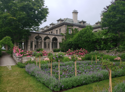 A historic mansion surrounded by vibrant flower gardens and lush greenery on a sunny day.