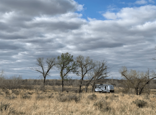 A camper parked among sparse trees in a grassy landscape under a cloudy sky.