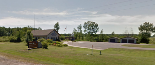 A small municipal building surrounded by greenery, with a parking lot and a sign for Pine Lake.
