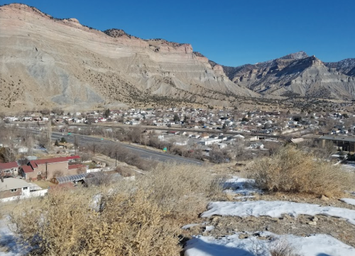 A panoramic view of a mountainous landscape with a small town and highway below, under a clear blue sky.