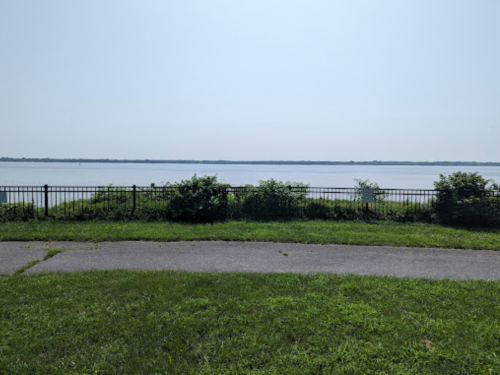 A calm river view with a grassy area in the foreground and a fence along the water's edge under a clear sky.