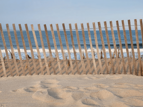 A wooden fence stands on a sandy beach, with ocean waves and a clear sky in the background.