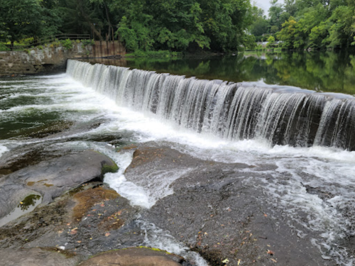 A serene waterfall cascades over a stone ledge into a calm river, surrounded by lush green trees.