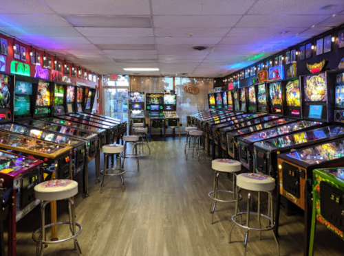 A vibrant arcade room filled with rows of pinball machines and colorful lights, featuring stools for players.