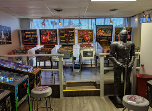 A retro arcade with pinball machines and a knight statue, featuring colorful lights and a cozy atmosphere.