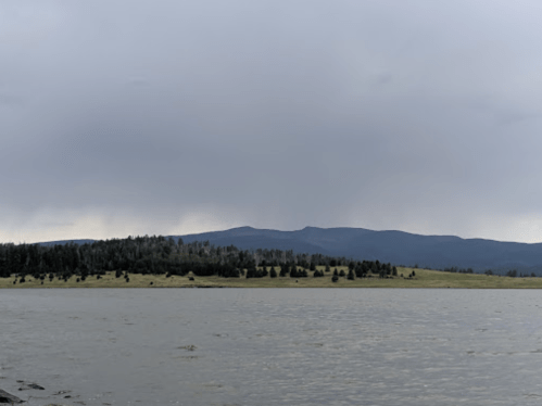A serene lake scene with a distant mountain range under a cloudy sky, surrounded by trees and grassy land.