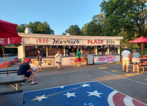 A busy outdoor eatery named Harry's Place, with patrons ordering food and red umbrellas providing shade.