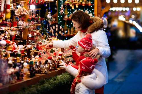 A woman and two children admire festive decorations at a holiday market, surrounded by colorful ornaments and lights.