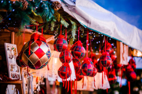 Colorful holiday ornaments with red ribbons hang from a festive market stall, adorned with greenery and lights.