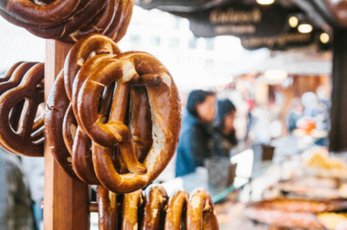 A display of freshly baked pretzels hanging at a market, with blurred shoppers in the background.