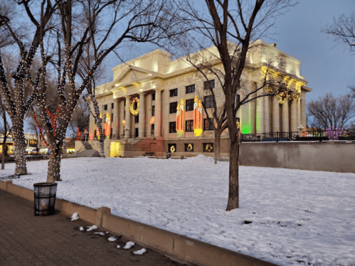 A snow-covered park with trees wrapped in lights, featuring a brightly lit building decorated for the holidays.