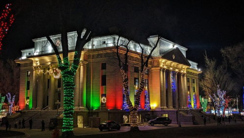 A historic building illuminated with colorful holiday lights at night, surrounded by decorated trees.