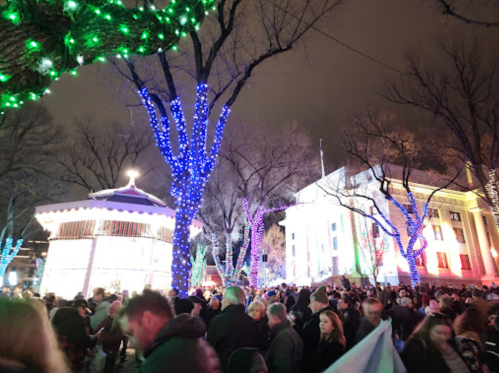 A festive crowd gathers at night, surrounded by colorful lights on trees and buildings, with a carousel in the background.