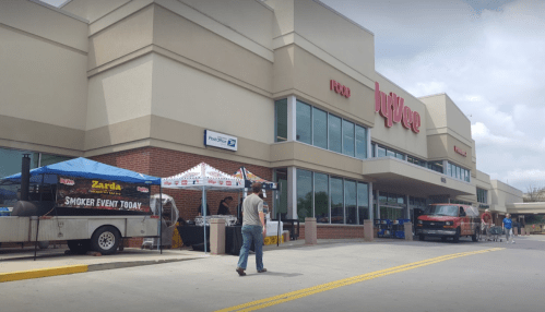 A grocery store exterior with tents for a smoker event, people milling about, and a truck parked nearby.