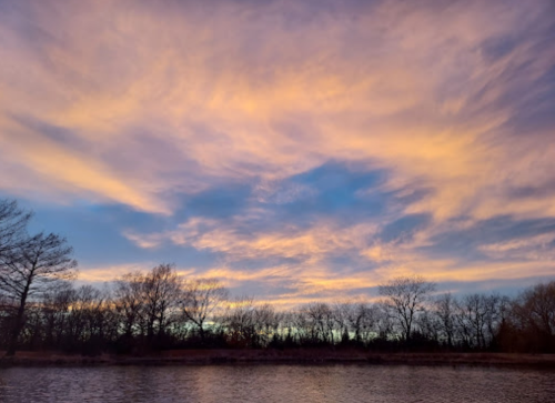 A serene sunset sky with colorful clouds above a calm river, framed by silhouetted trees along the shore.