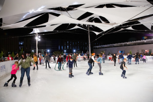 A lively ice skating rink at night, filled with people of all ages enjoying skating under a large canopy.