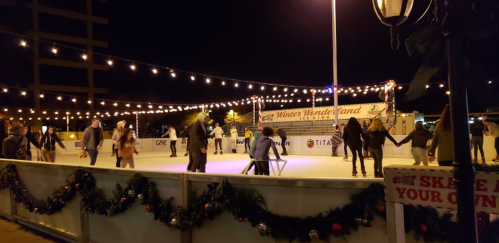 A lively ice skating rink at night, decorated with string lights and festive garlands, with people skating and enjoying the scene.