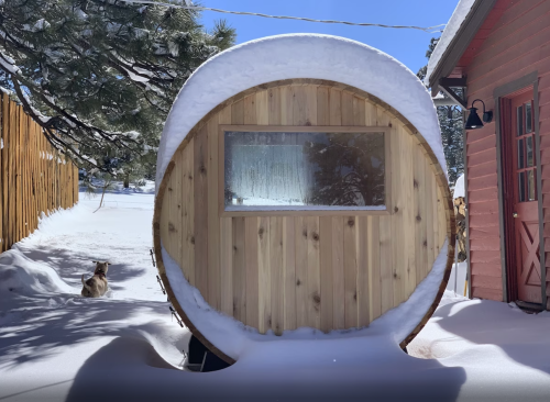 A wooden barrel-shaped structure covered in snow, with a window, beside a house and a cat sitting in the snow.