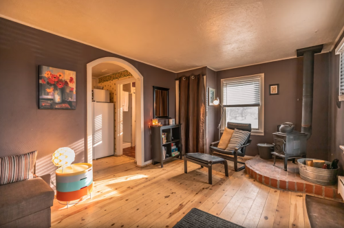 Cozy living room with wooden floors, a stove, and natural light, featuring a sofa, chair, and decorative elements.