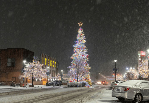 A brightly lit Christmas tree stands in a snowy street, surrounded by festive lights and falling snowflakes.