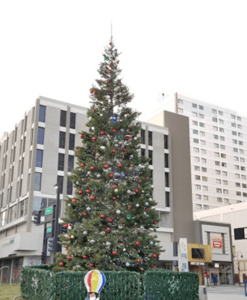 A tall Christmas tree decorated with colorful ornaments stands in an urban area near buildings.