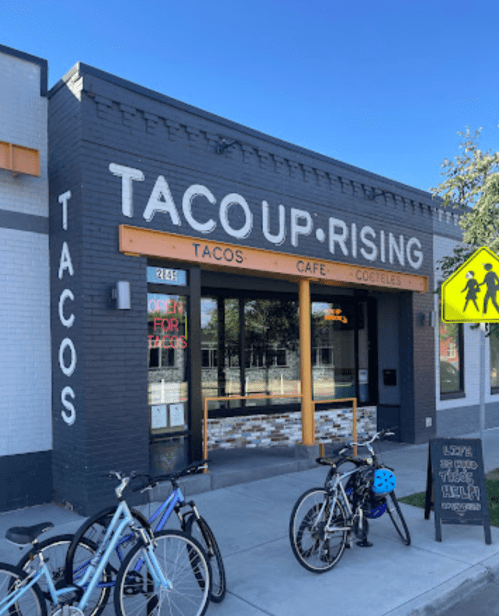 Exterior of Taco Up-Rising restaurant with bikes parked outside and a sign indicating it's open for tacos and cocktails.