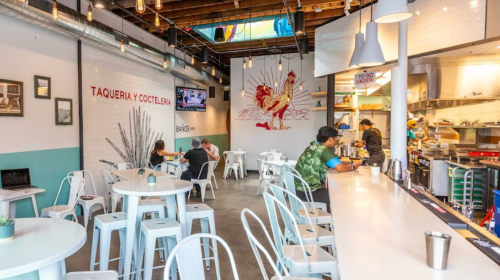 A modern taqueria with white tables, a vibrant mural, and patrons enjoying their meals in a casual setting.