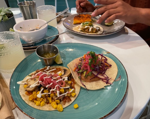 A plate with two tacos topped with various ingredients, a side of sauce, and a lime wedge, with food in the background.