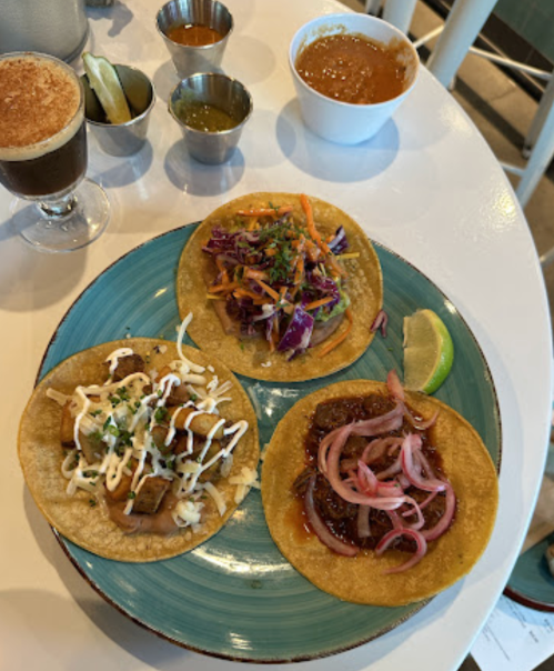 Three colorful tacos on a blue plate, accompanied by sauces and lime wedges on a white table.