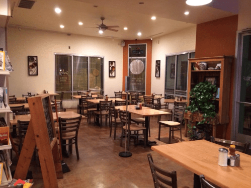 A spacious, empty restaurant with wooden tables and chairs, large windows, and warm lighting.
