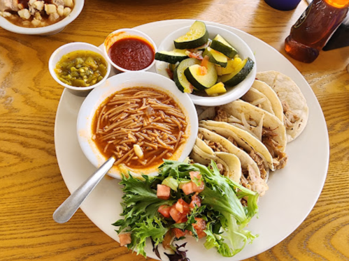 A plate of tacos with shredded meat, a side of soup, salad, and mixed vegetables, accompanied by sauces.