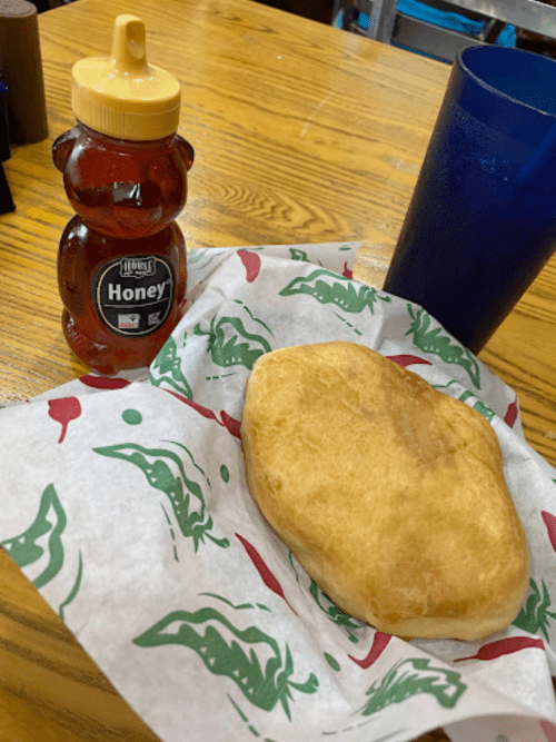 A honey bear bottle next to a golden-brown fried dough on a patterned paper, with a blue cup in the background.