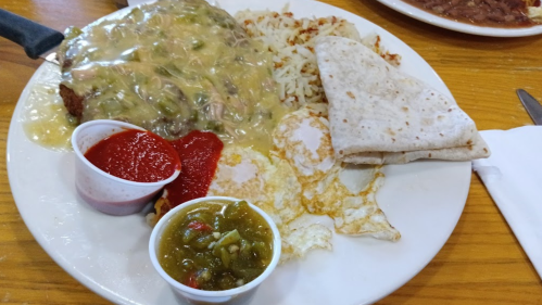 A plate of chicken smothered in green sauce, served with hash browns, eggs, tortillas, and two dipping sauces.
