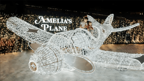Two children sit in a large, illuminated airplane sculpture with "Amelia's Plane" sign in a festive setting.