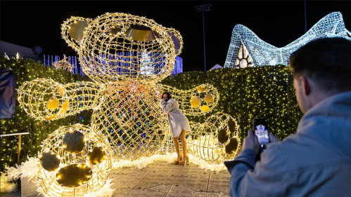 A person poses next to a large illuminated teddy bear display at a festive outdoor event.